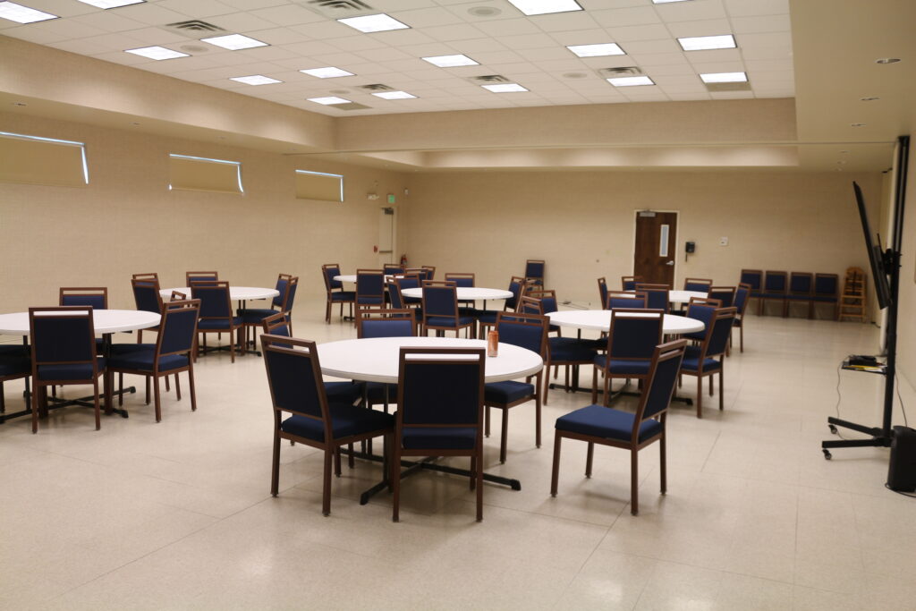 Photograph of multipurpose room. There are several tables each surrounded with chairs.